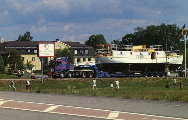Äldre passagerarbåt lastad på en låg trailer bakom en lastbil